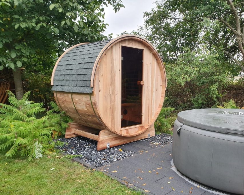 Holzfass-Sauna mit Eingang, umgeben von Bäumen und einer runden Badewanne.